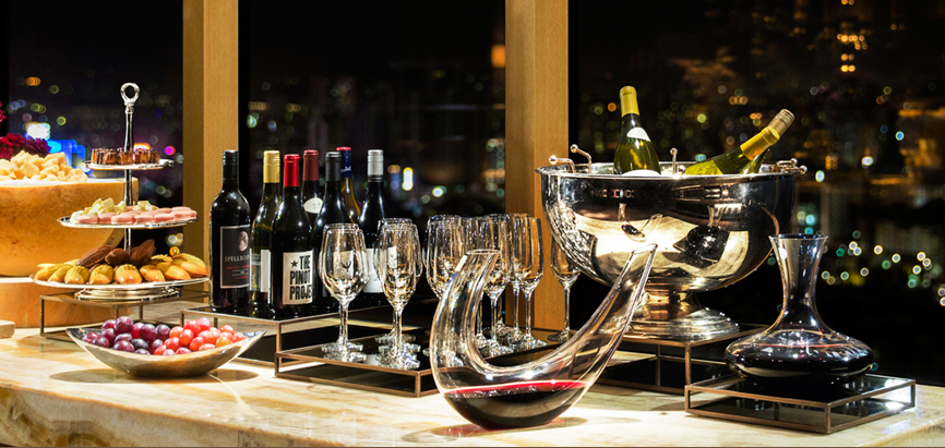 윈터 셀레브레이션 파티 혜택을 보여주는 이미지. 더 이그제큐티브 라운지 테이블 위에 여러 병의 와인과 와인 잔이 세팅되어 있다. 왼편에는 디저트 류가 담긴 3단 트레이와 과일이 담긴 접시가 놓여 있으며, 오른편에는 와인이 담긴 디켄터 2개 및 와인이 여러 병 담긴 아이스 버킷이 놓여 있다. 뒤편으로는 도심 야경이 펼쳐진다.