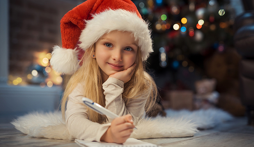 집 안의 풍경, 뒤에는 크리스마스 장식이 있고 산타 모자를 쓴 어린이가 펜을 들고 웃으면서 정면을 보고 있는 사진