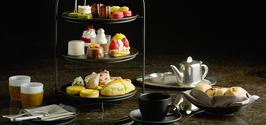 더 라이브러리 애프터눈 티 세트를 보여주는 이미지. 테이블 위에 놓인 3단 트레이에 케이크, 마카롱 등 디저트와 세이버리가 차례로 올려져 있다. 오른편에는 주전자와 찻잔, 스콘이 담긴 접시가 차례로 놓여 있다.
