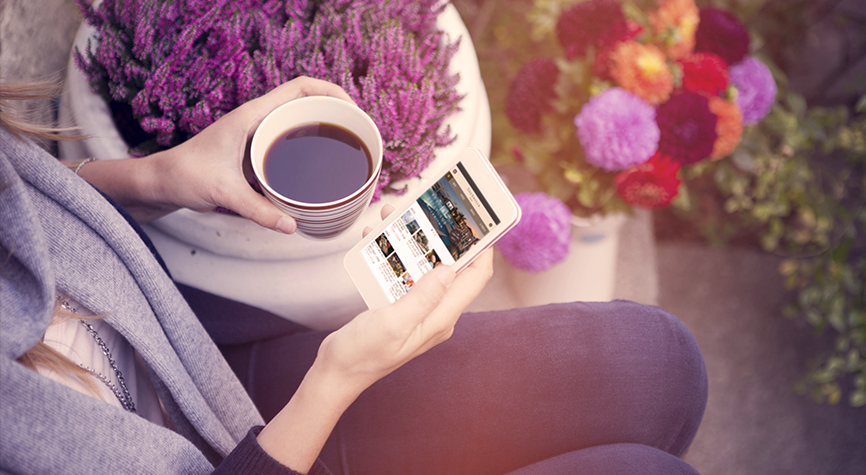 여성이 왼손에 커피가 든 잔을 들고 오른손에는 제주신라호텔 모바일 메인 화면이 떠 있는 휴대전화를 들고 있으며 주변에는 다양하고 예쁜 꽃들이 있는 사진