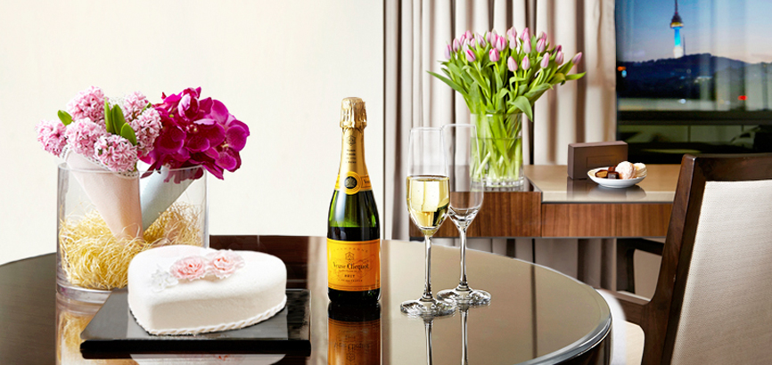 首尔新罗酒店客房的桌子上放着黄牌香槟一瓶，两个香槟杯，水果奶油蛋糕和雪白的花束装点。