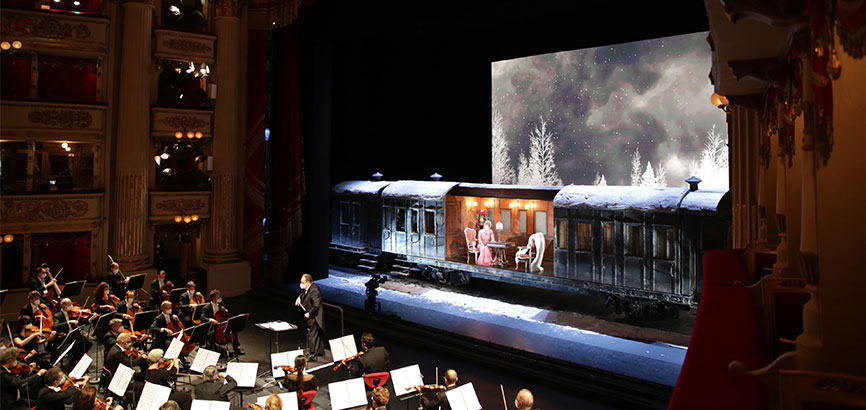 라스칼라 갈라 콘서트의 전경을 보여준다. 왼쪽에는 오케스트라, 오른쪽에는 오페라 무대 위 성악가가 노래를 부르고 있다. 