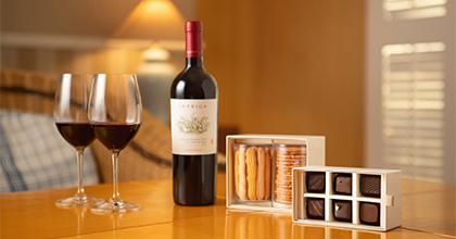 테이블 위 와인이 담겨진 와인 잔2개, 하우스 와인 1병, 쿠키 2종과 초콜렛 6개가 있는 이미지