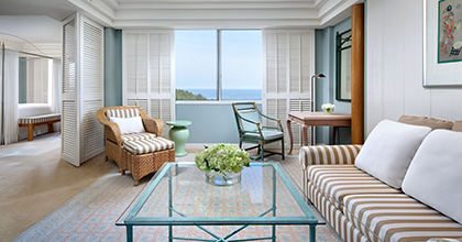 Image of a corner suite living room with pastel-toned interiors, a sofa for two, a table, rattan chairs, and a dining table overlooking the sea.