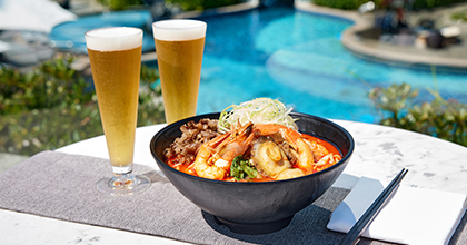 Image of a white table with jjamppong topped with Korean beef, shrimp, and abalone, alongside two beers, with a pool in the background