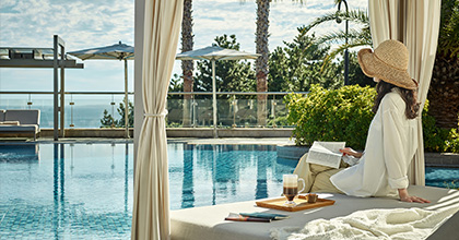 하얀 모자를 쓴 여성이 워터 베드에 기대어 수영장을 바라보며 책을 읽고 있는 이미지.