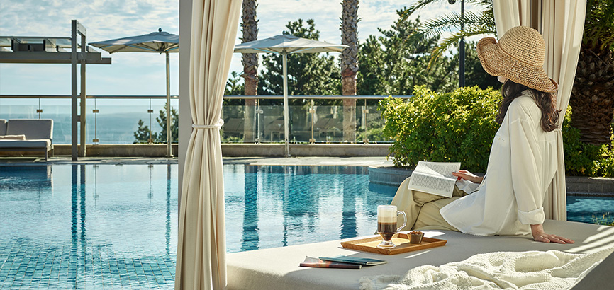 하얀 모자를 쓴 여성이 워터 베드에 기대어 수영장을 바라보며 책을 읽고 있는 이미지.